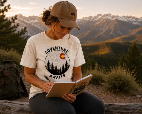 Woman wearing Adventure Awaits Colorado T-Shirt journaling on a mountain overlook