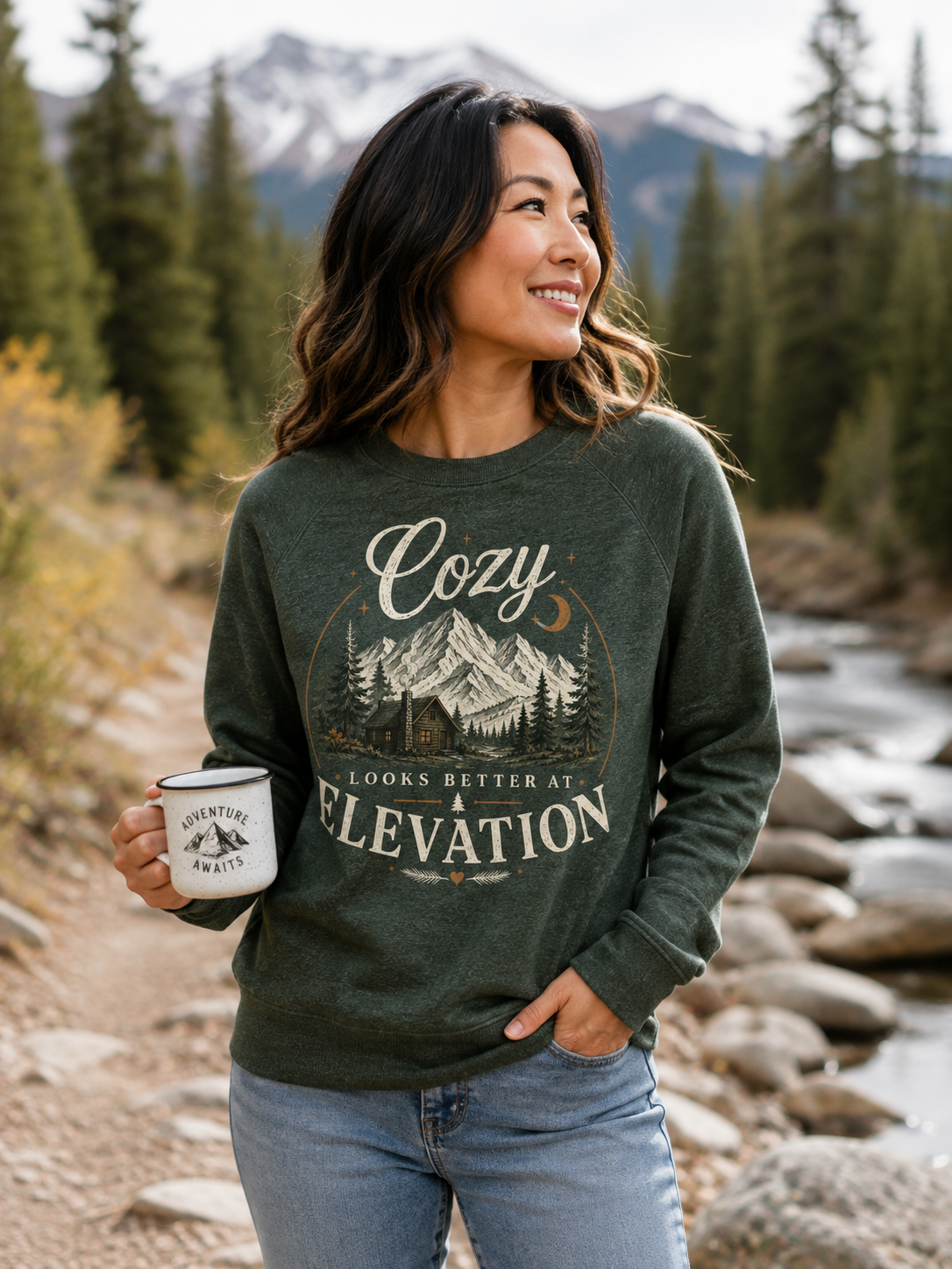 Woman wearing Cozy Looks Better at Elevation sweatshirt by a Colorado mountain stream holding a camp mug