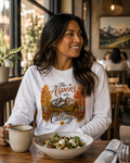 A woman sitting at a wooden table in a cozy cafe setting, wearing a white long-sleeve graphic t-shirt with 'The Aspen are Calling' and an autumn mountain design. She is smiling and looking to the side, with a bowl of salad and a mug in front of her.