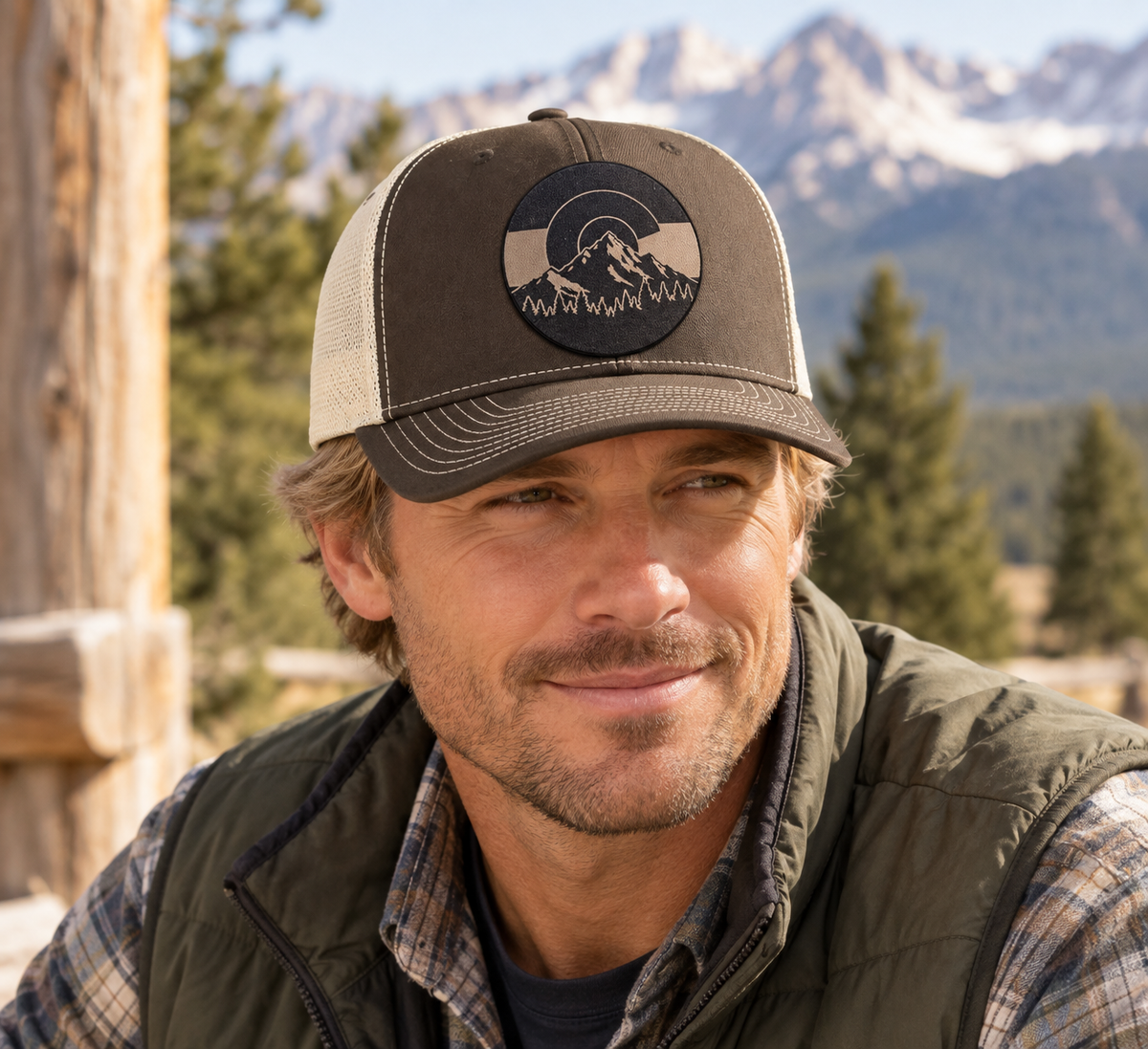 Person wearing a two-tone Mountain Heritage trucker hat with a leather mountain patch against a Colorado mountain backdrop