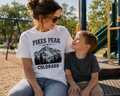 A woman and a young boy sitting together at a playground, both wearing matching graphic t-shirts. The woman's white t-shirt features a black graphic of a mountain and pine trees with the text 'PIKES PEAK COLORADO'.