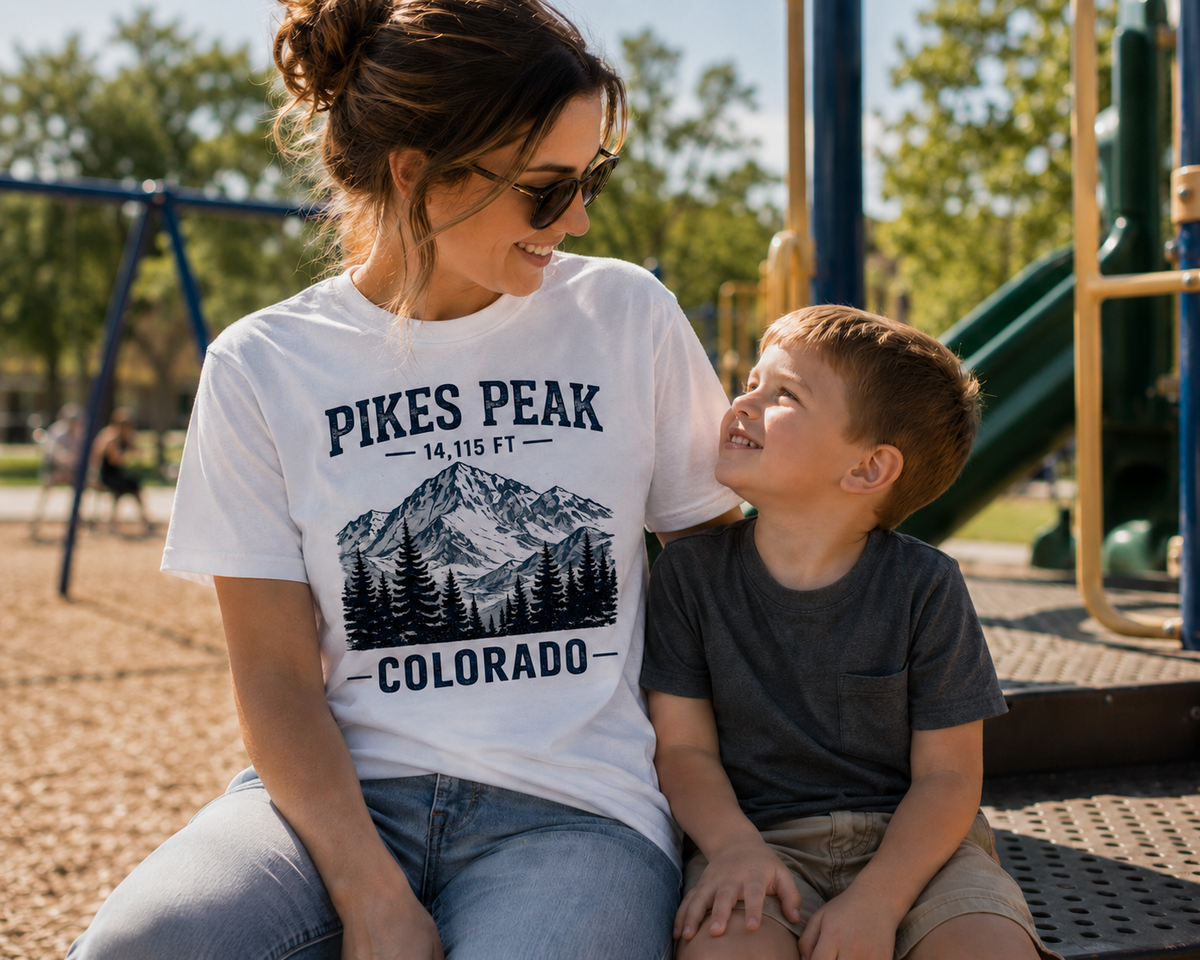 A woman and a young boy sitting together at a playground, both wearing matching graphic t-shirts. The woman's white t-shirt features a black graphic of a mountain and pine trees with the text 'PIKES PEAK COLORADO'.