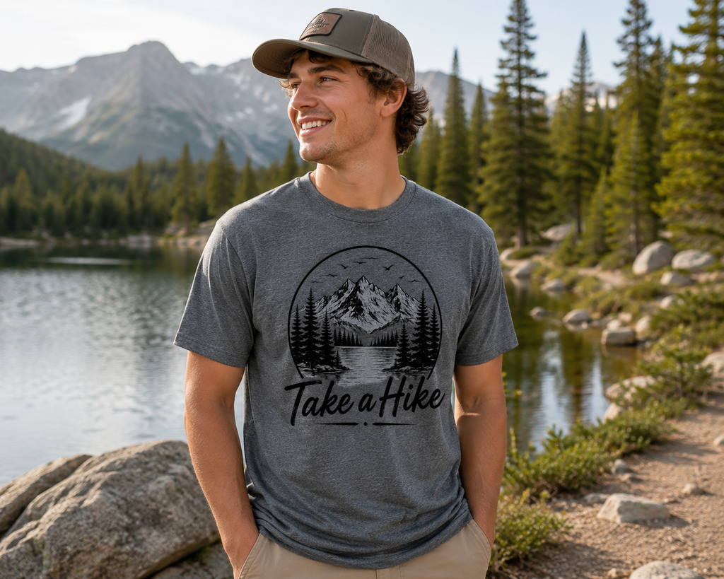 A man wearing a heather gray t-shirt with a black 'Take a Hike' mountain graphic, standing outdoors in front of a scenic mountain lake.